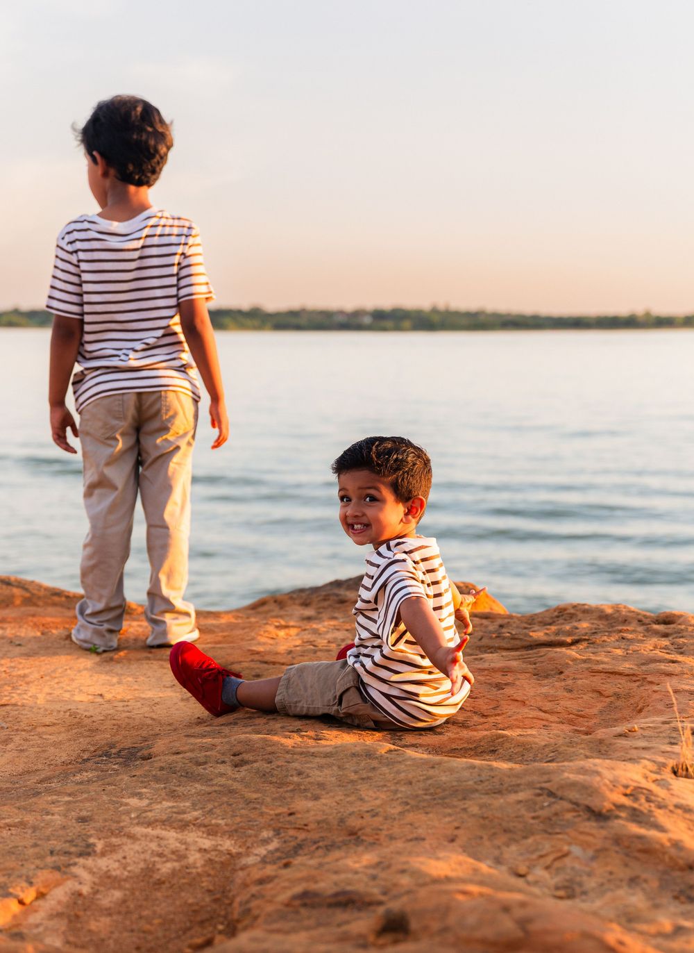Two young boys sitting and standing on rocky shore by calm lake at golden sunset hour.