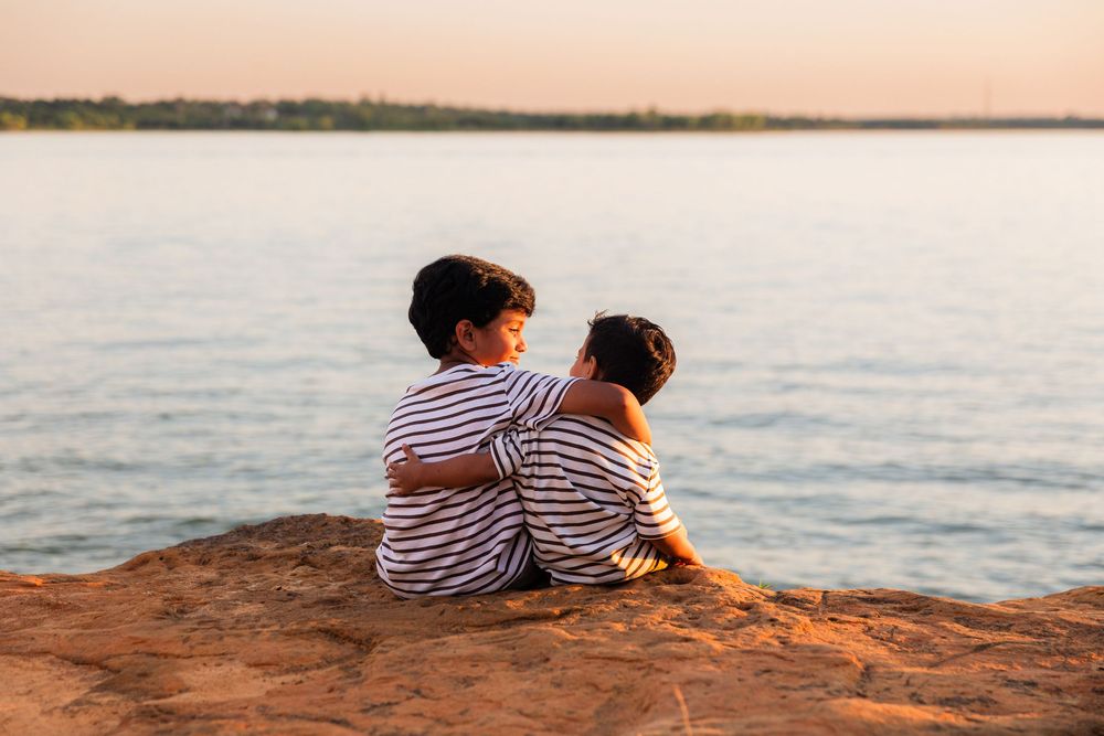 Two children in striped shirts sit embracing on a rock overlooking a calm lake at sunset.