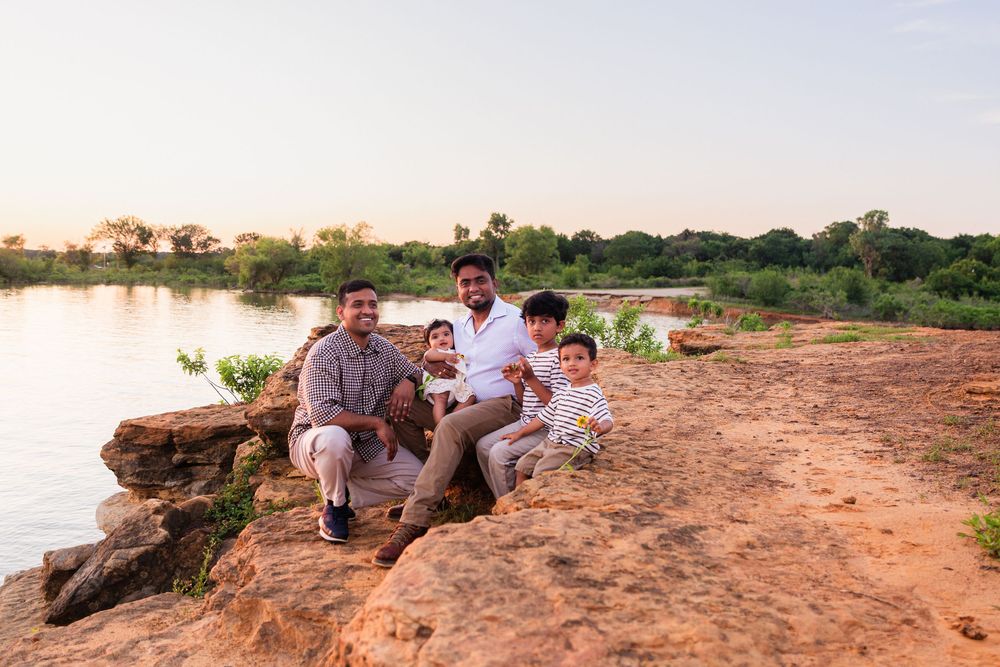 Family of five posing on rocky riverside at sunset with lush green trees and calm water in background.