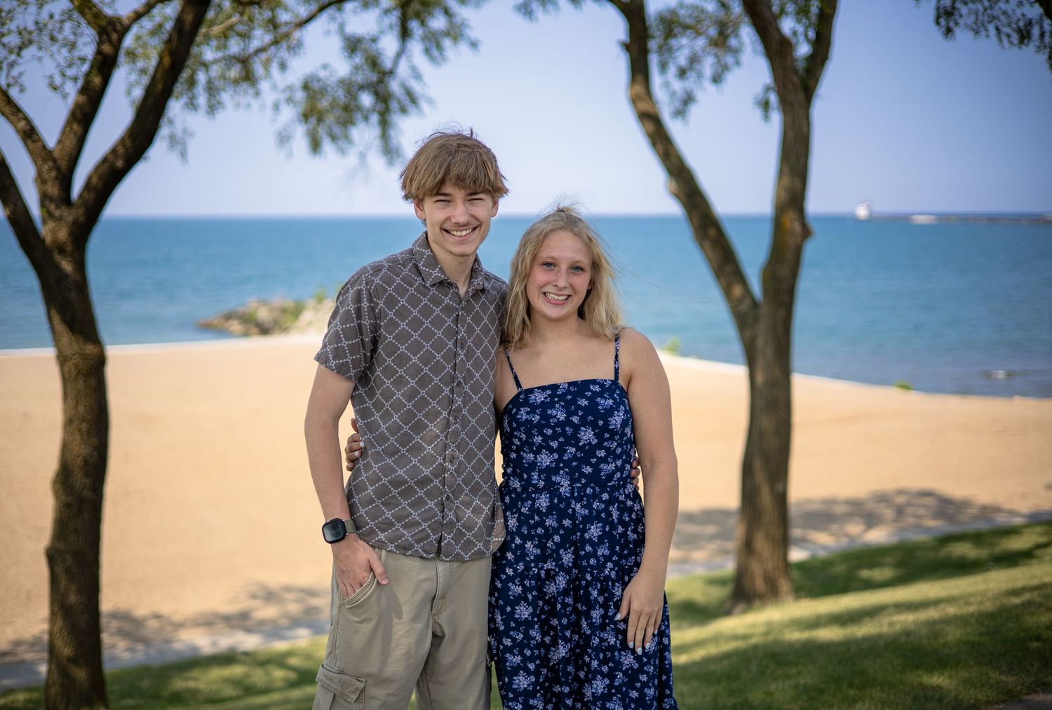 Smiling teenage boy and blonde girl posing together under trees with a sunny beach in the background.