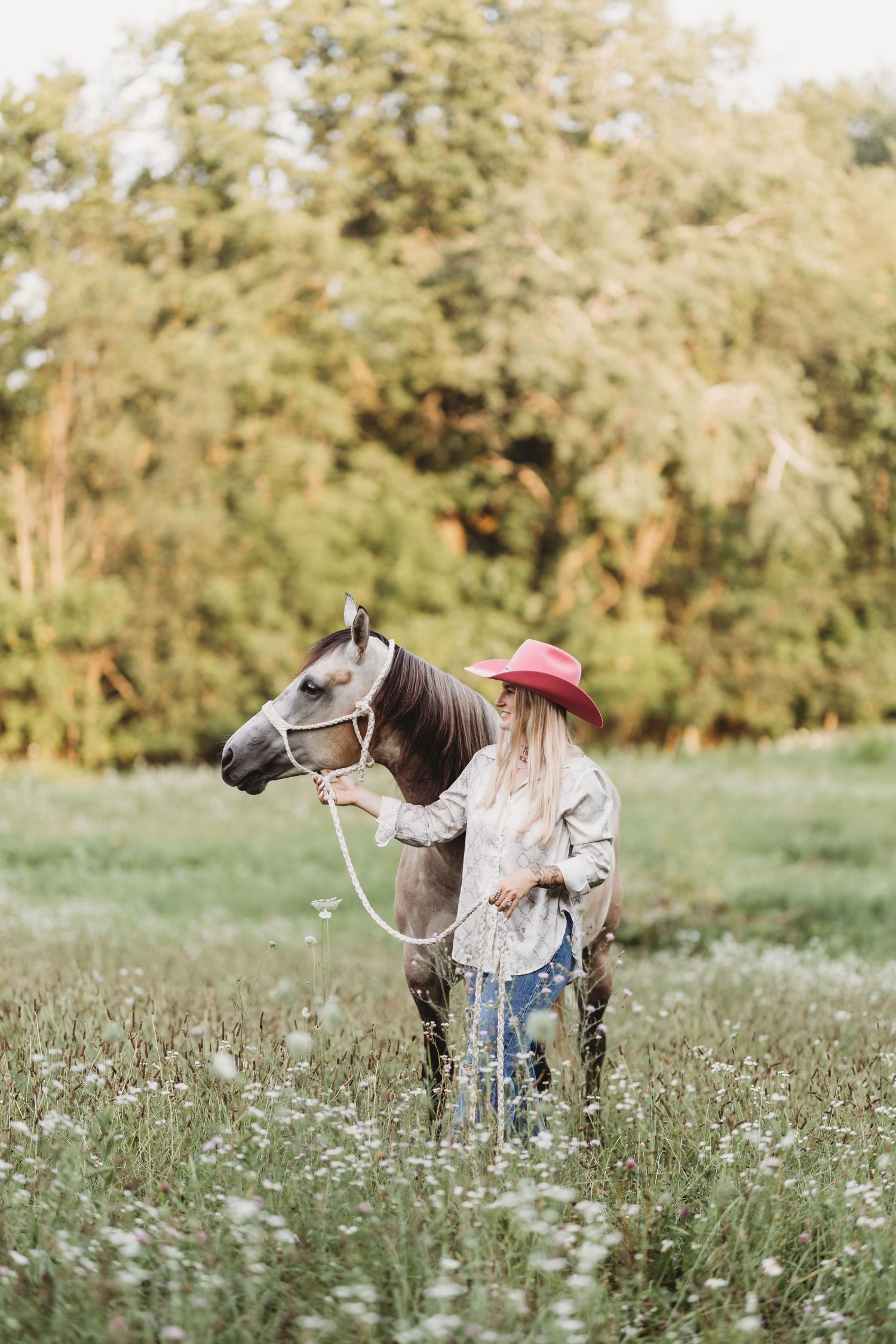 Woman in pink cowboy hat leading a brown horse through a wildflower meadow with green trees in background.