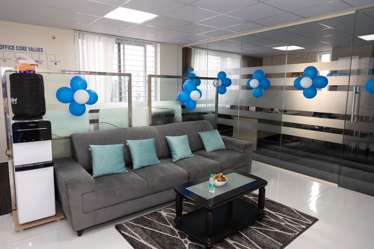 Modern office lounge with gray sofa, teal pillows, blue flower decorations, glass partitions, and a water cooler on white flooring.