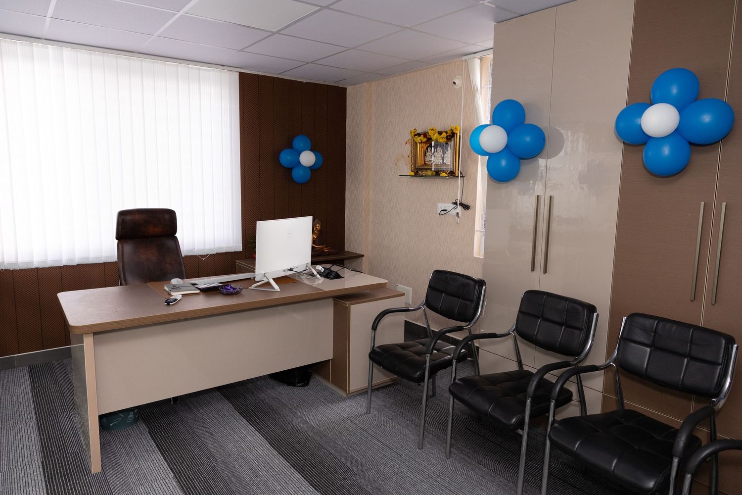 Modern office reception area with a wooden desk, computer, black chairs, and blue balloon flower decorations on the walls.