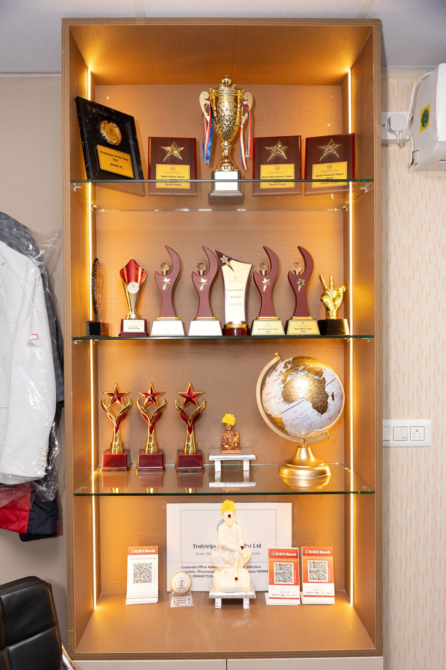 Illuminated display cabinet showcasing multiple gold and red trophies, awards, and a decorative globe on glass shelves.