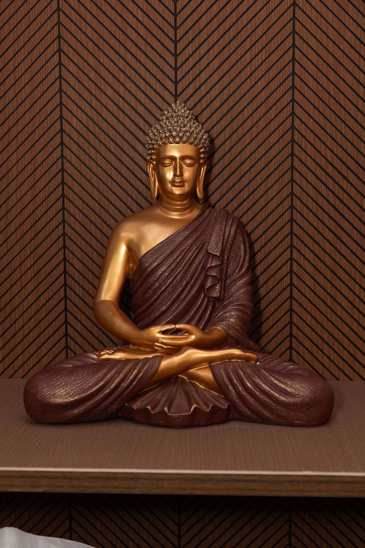 Golden Buddha statue in meditation pose on wooden shelf with decorative chevron wall background.