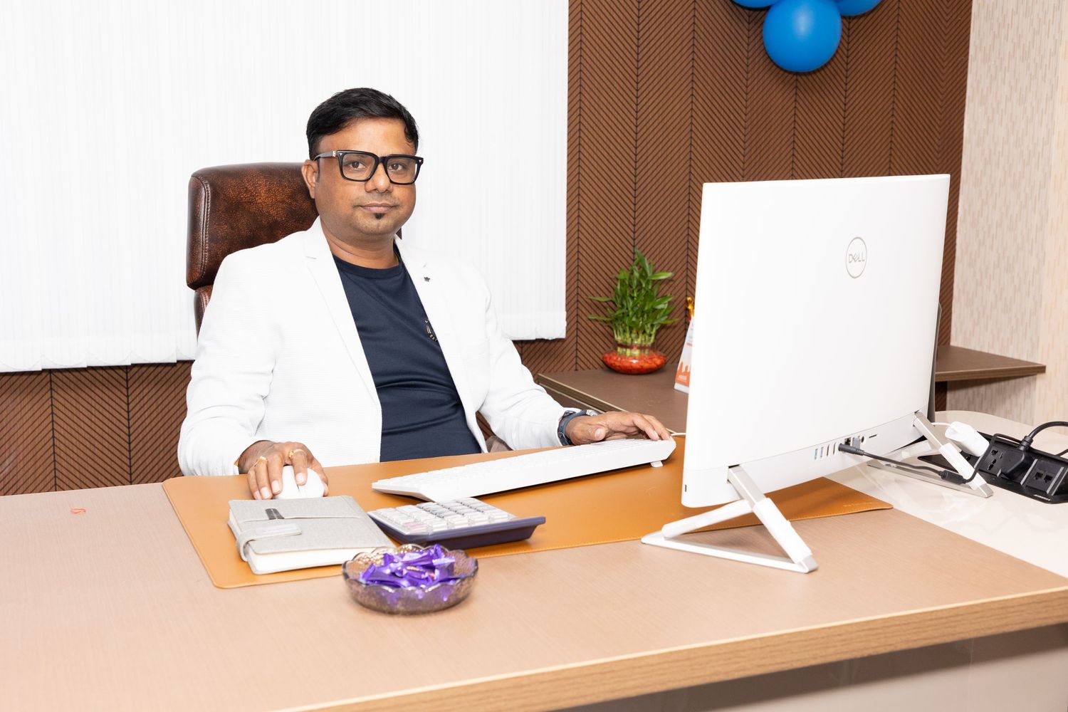 Professional man in white blazer sitting at office desk with computer, calculator, and plant in modern workspace.