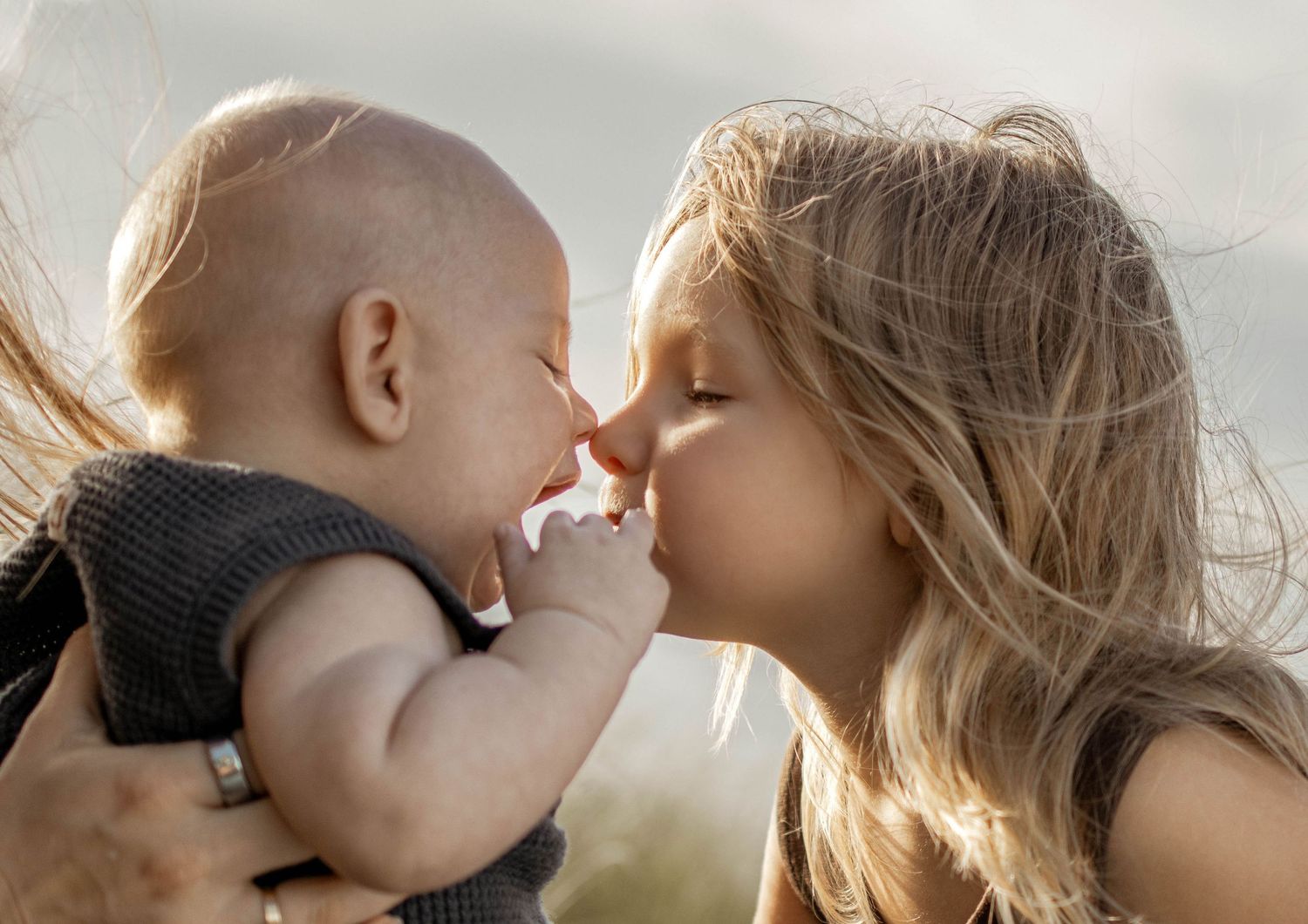 A young blonde girl tenderly touches noses with a baby in a heartwarming outdoor moment.