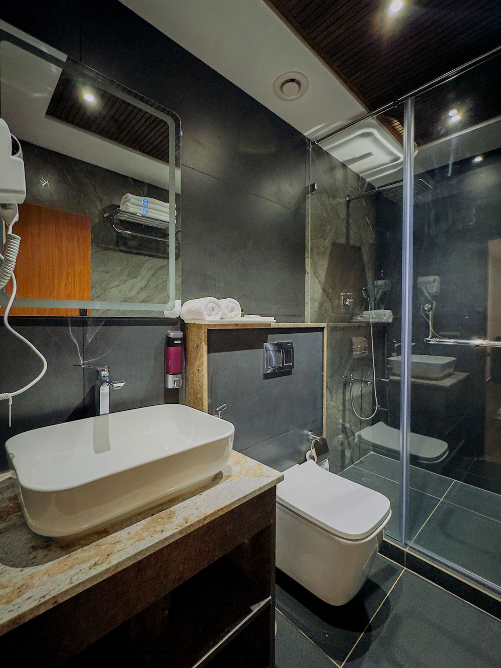 Modern hotel bathroom with vessel sink, marble vanity, glass shower enclosure, and dark slate walls.