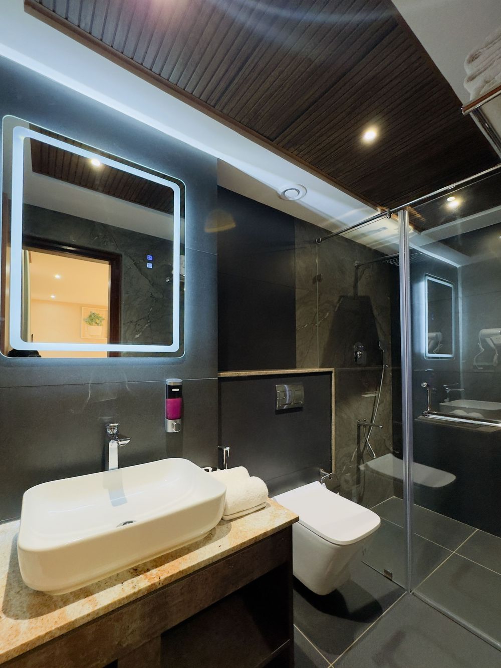Modern hotel bathroom with LED mirror, vessel sink, wall-mounted toilet, and glass shower enclosure with dark marble tiles.