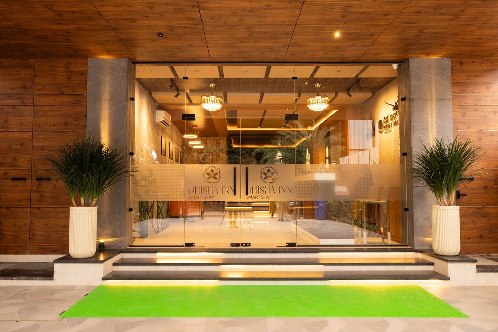 Modern hotel entrance with glass doors, wood walls, green carpet, and tropical plants in elegant lobby.