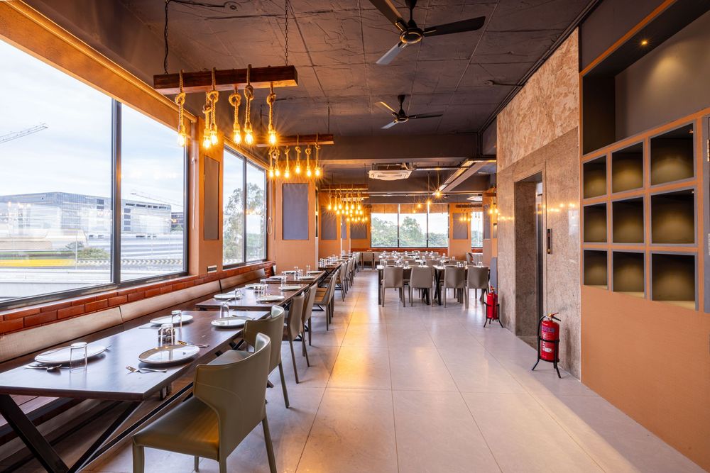 Modern restaurant interior with pendant lights, set dining tables, large windows, and wall shelving in warm neutral tones.