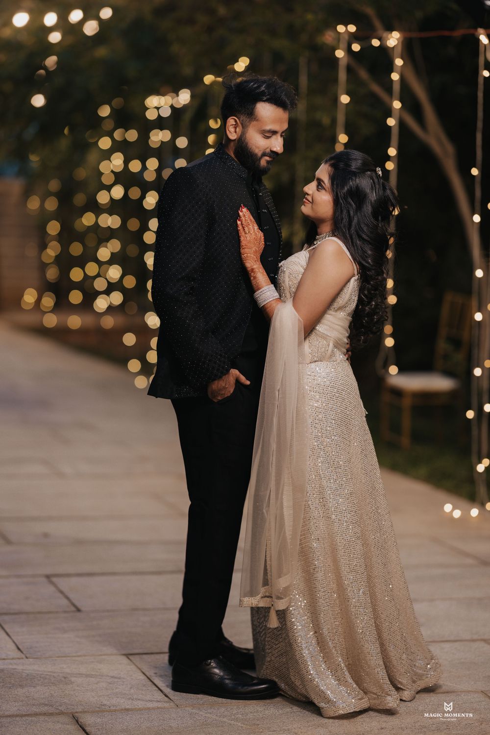 Couple in elegant attire sharing a romantic moment outdoors, surrounded by warm bokeh string lights at night.