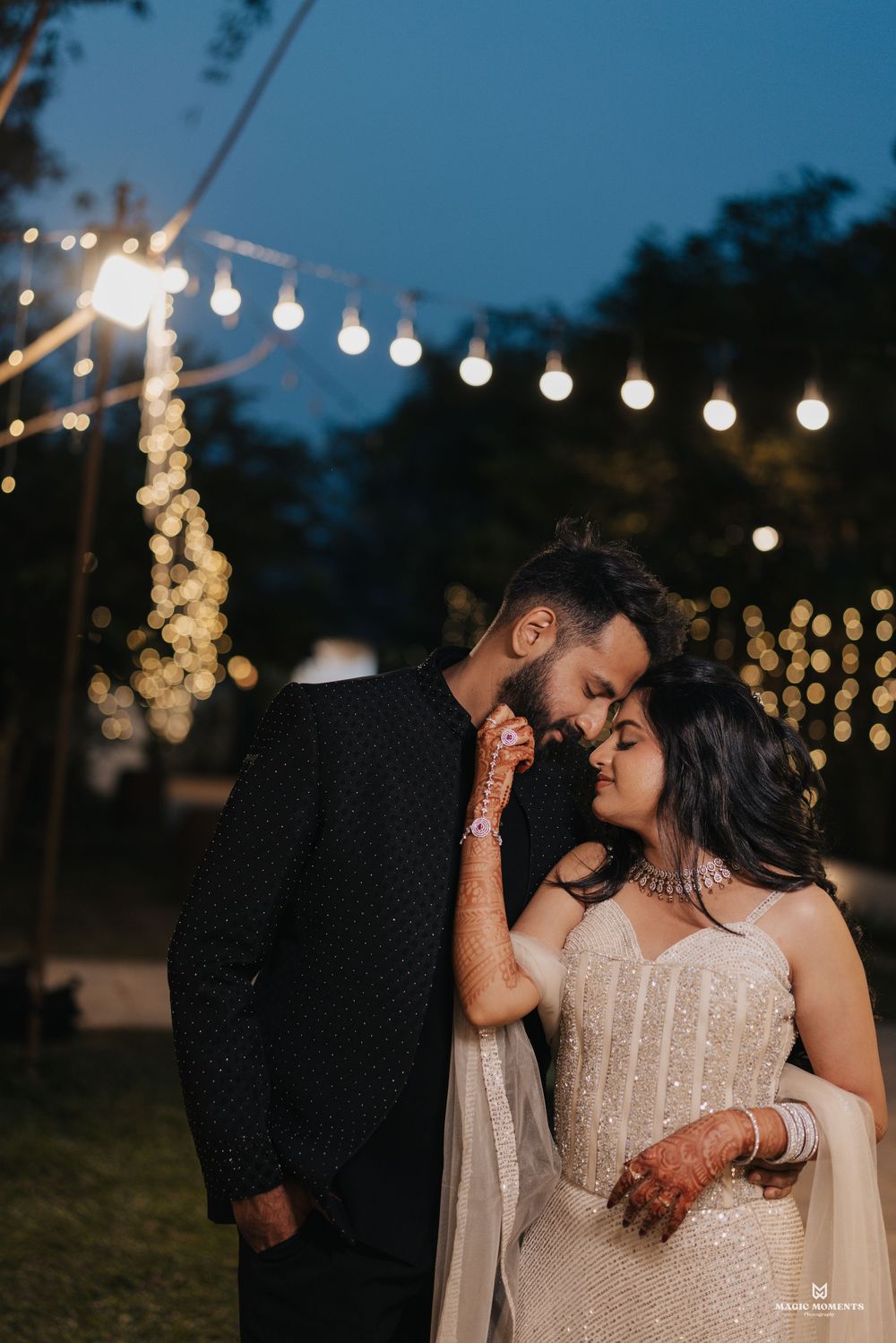 Romantic couple in elegant wedding attire sharing an intimate moment under string lights at a twilight outdoor celebration.