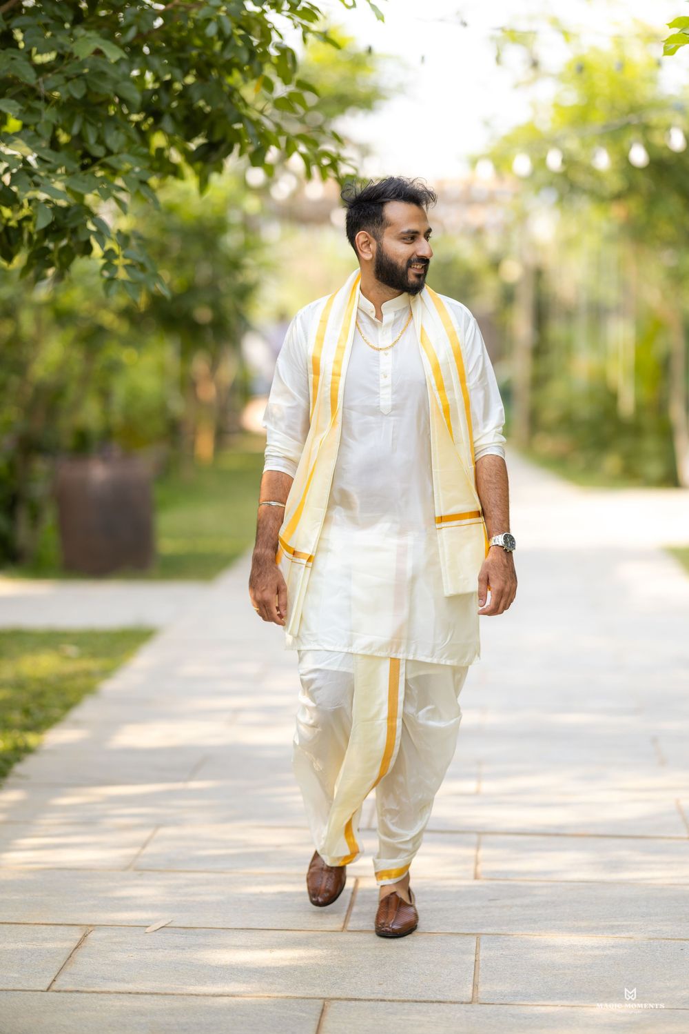 Man in white kurta and dhoti with yellow-bordered shawl walking on a garden pathway.