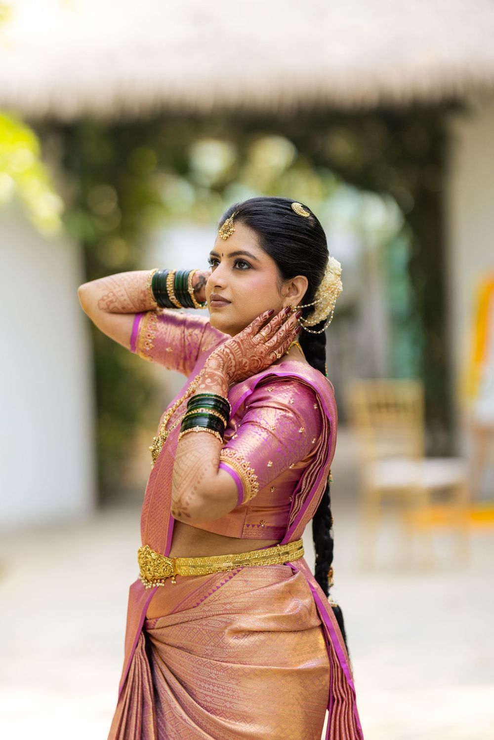 Indian bride in pink silk saree with gold jewelry and mehndi, posing gracefully outdoors.