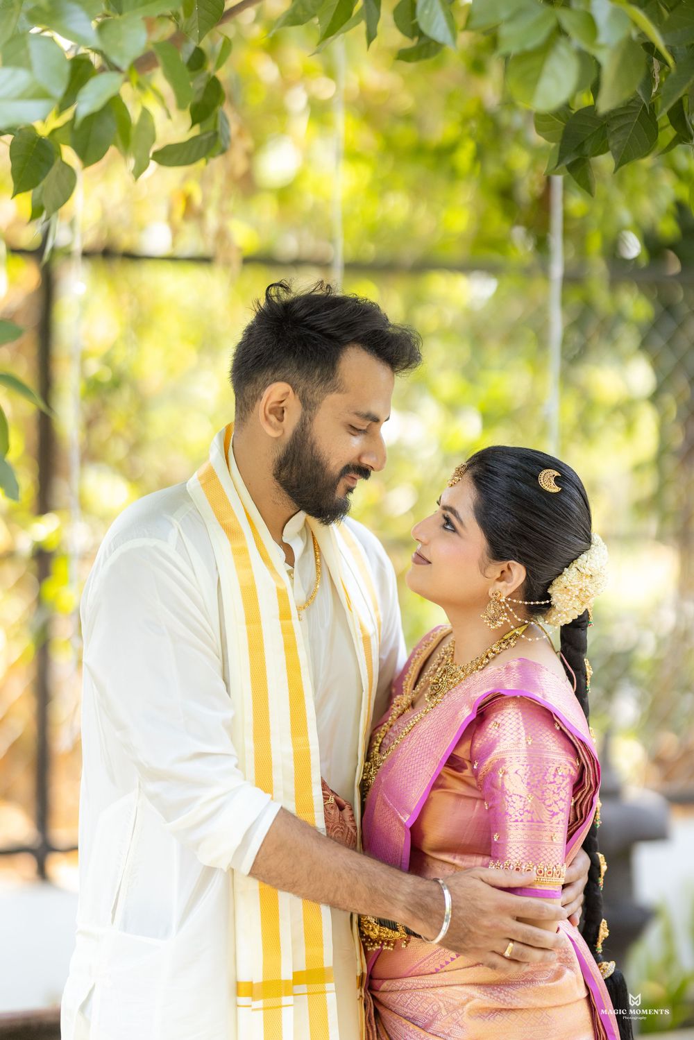 South Indian couple in traditional wedding attire, groom in white kurta, bride in pink silk saree with gold jewelry.