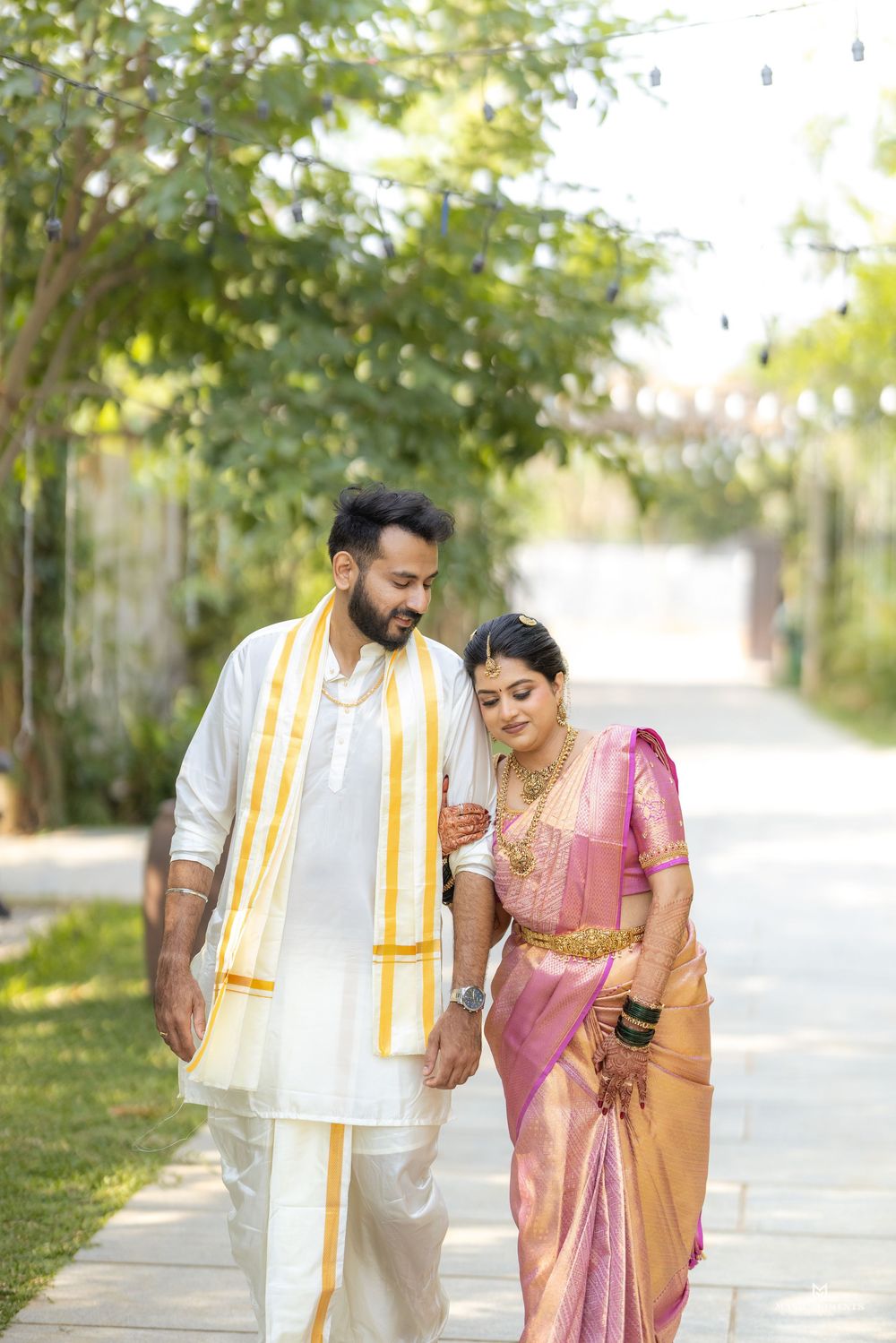 Indian couple in traditional wedding attire walking together, man in white dhoti and woman in pink silk saree with gold jewelry.