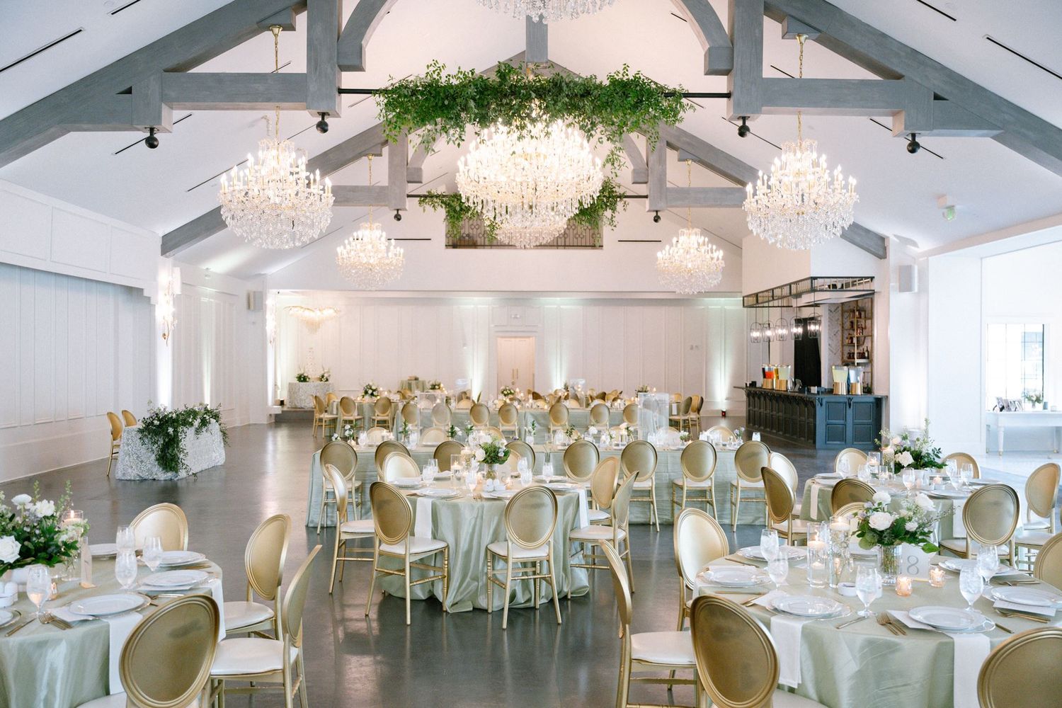 Elegant wedding reception hall with crystal chandeliers, greenery ceiling installation, round tables with white linens, and gold chairs.