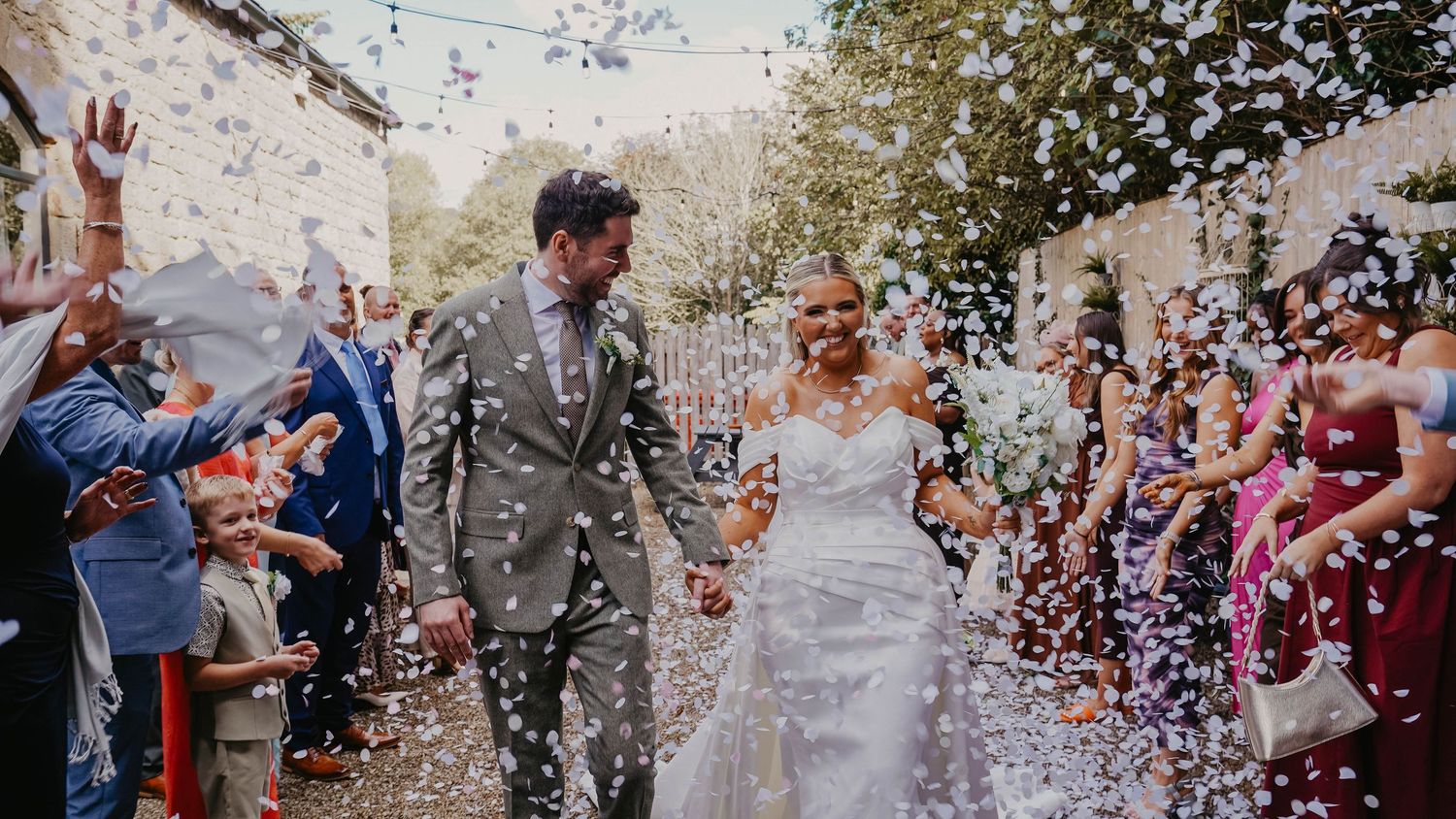 Bride and groom walk through colorful confetti shower surrounded by cheering wedding guests outdoors.