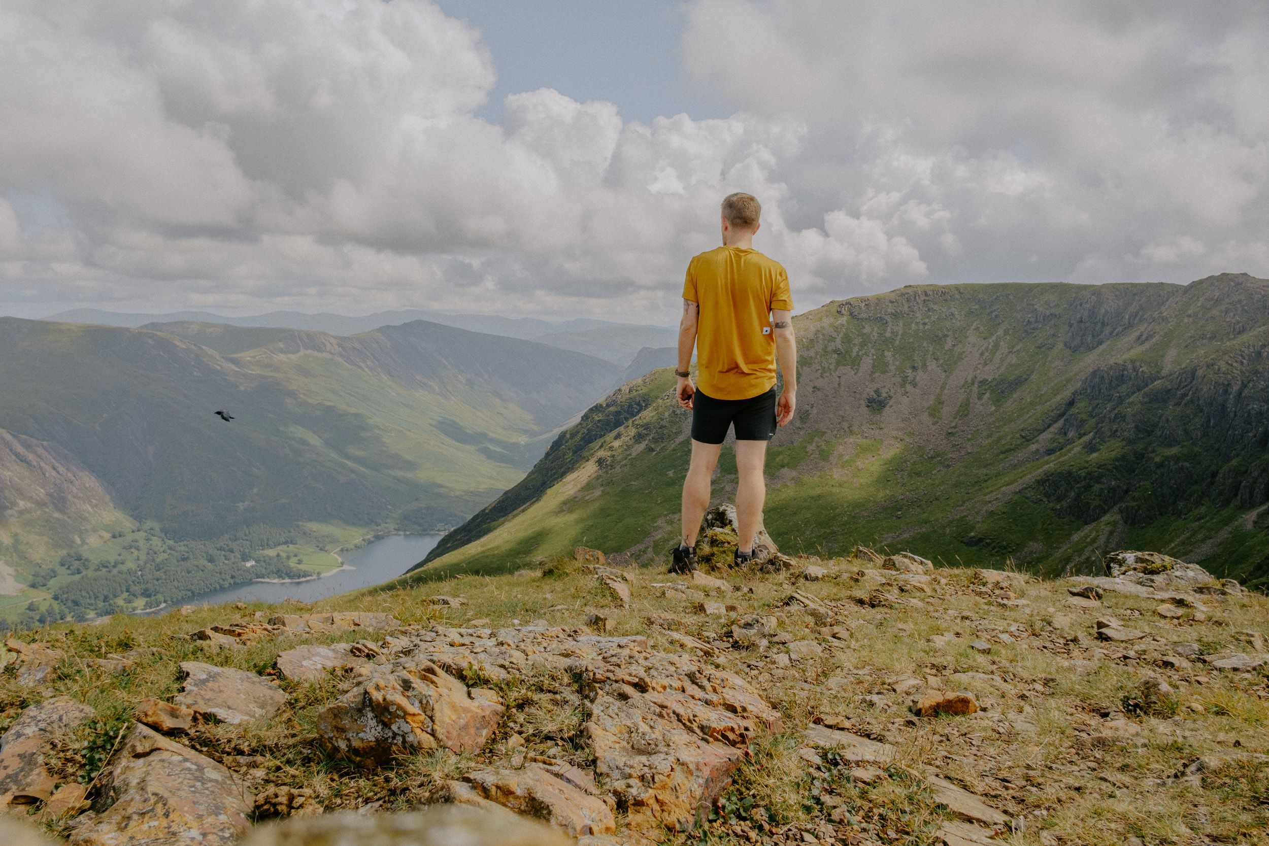 Hiker in yellow shirt stands on rocky mountain summit overlooking a scenic green valley and lake under cloudy skies.
