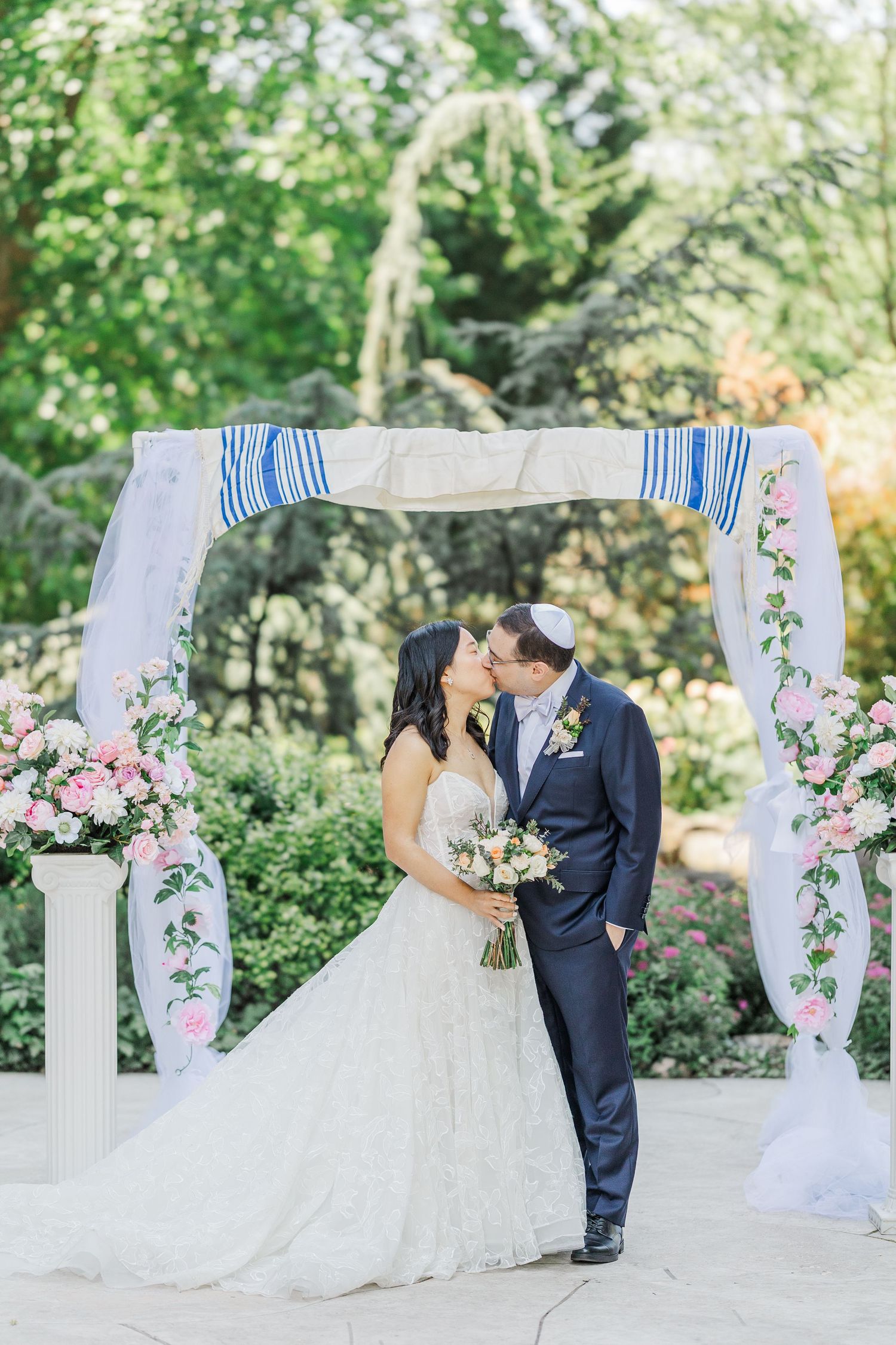 Bride and groom share a first kiss under a floral chuppah with pink roses and white draping at an outdoor Jewish wedding.