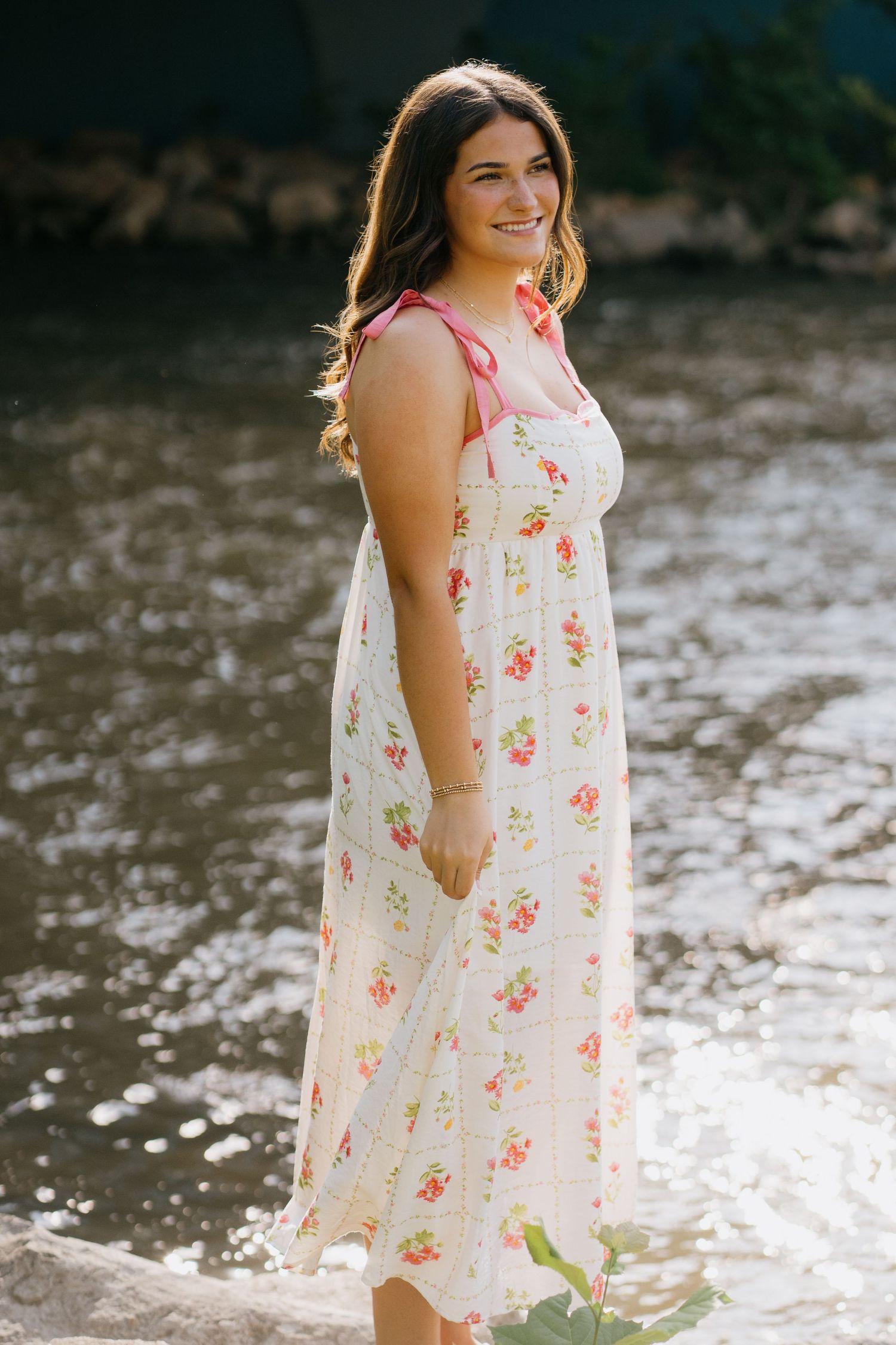 Smiling young woman in a white floral midi dress with pink tie straps standing near a sunlit river.