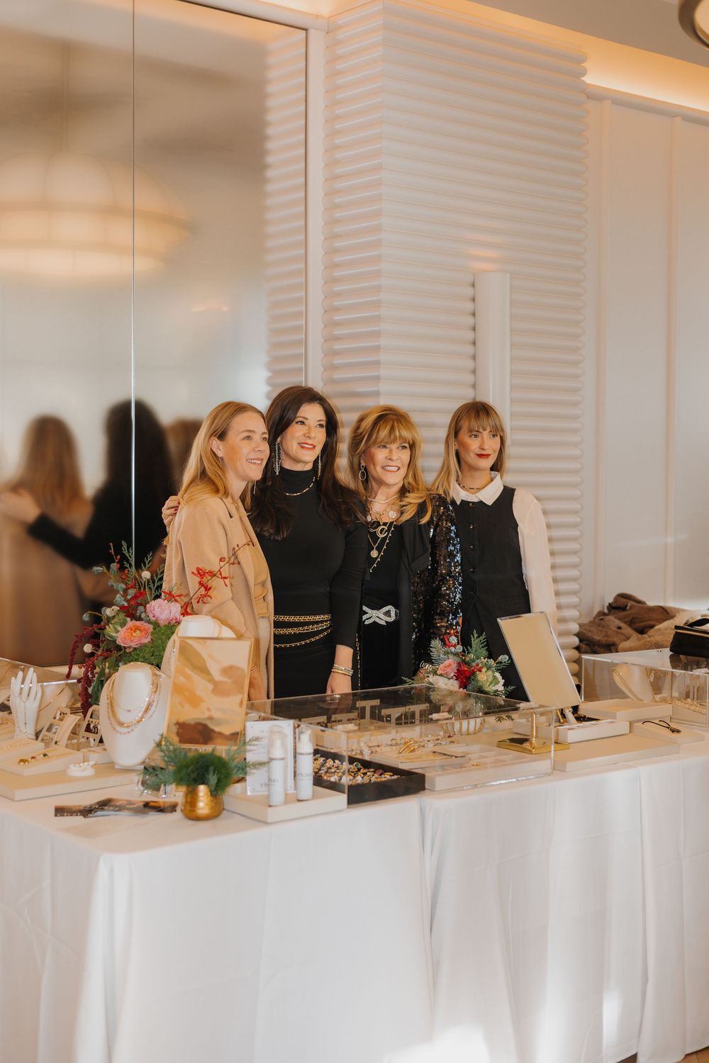 Four women smiling behind a jewelry display table at an elegant indoor market event with white decor.