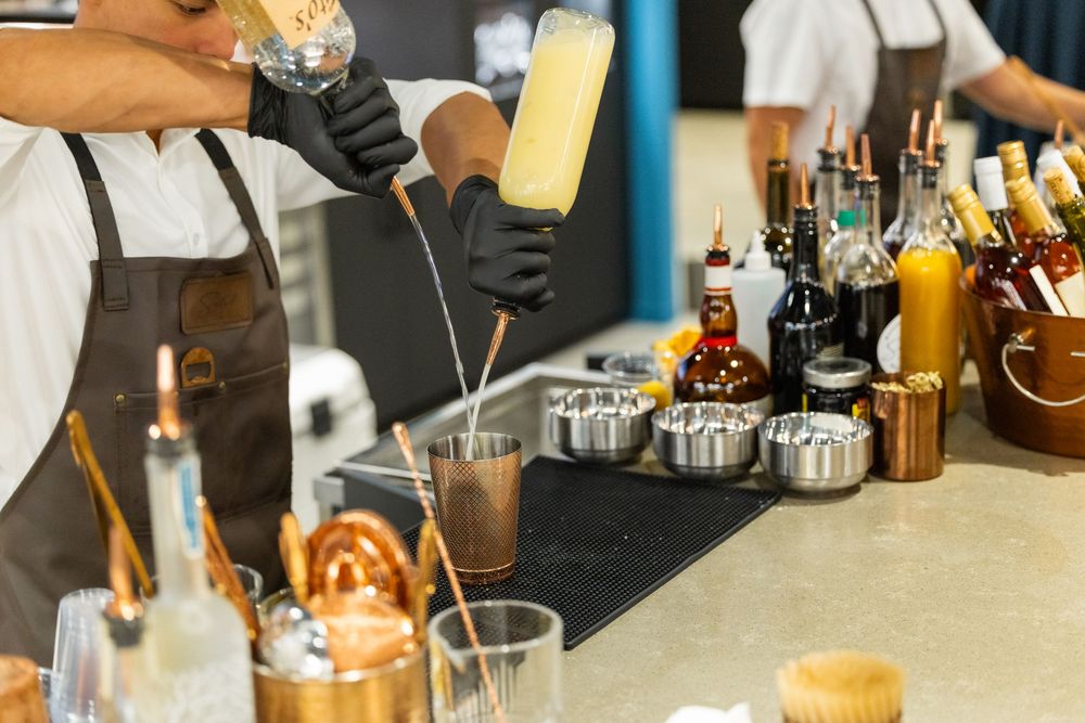 Bartender in black gloves pouring cocktail shaker at a bar with bottles and metal cups on counter.