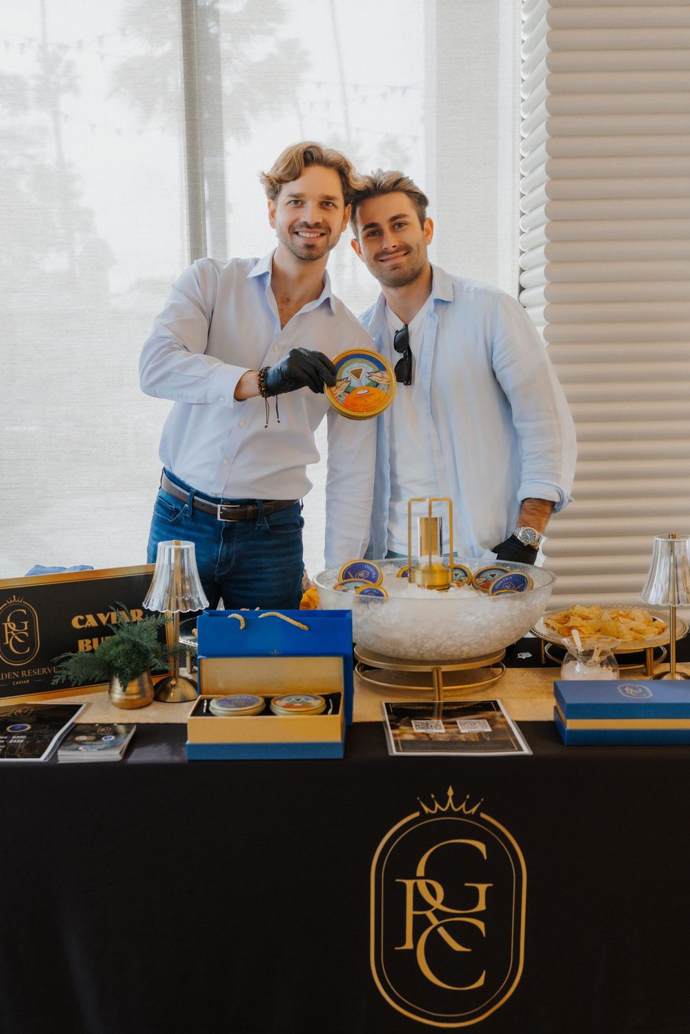 Two men smile behind a caviar display table with a luxury brand logo at an upscale catering event.