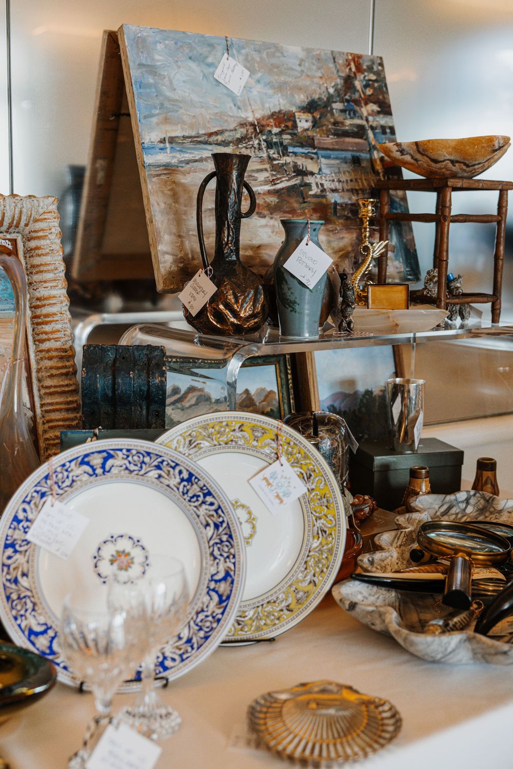 Antique shop display with decorative plates, crystal glass, vases, and a coastal painting on a cluttered table.