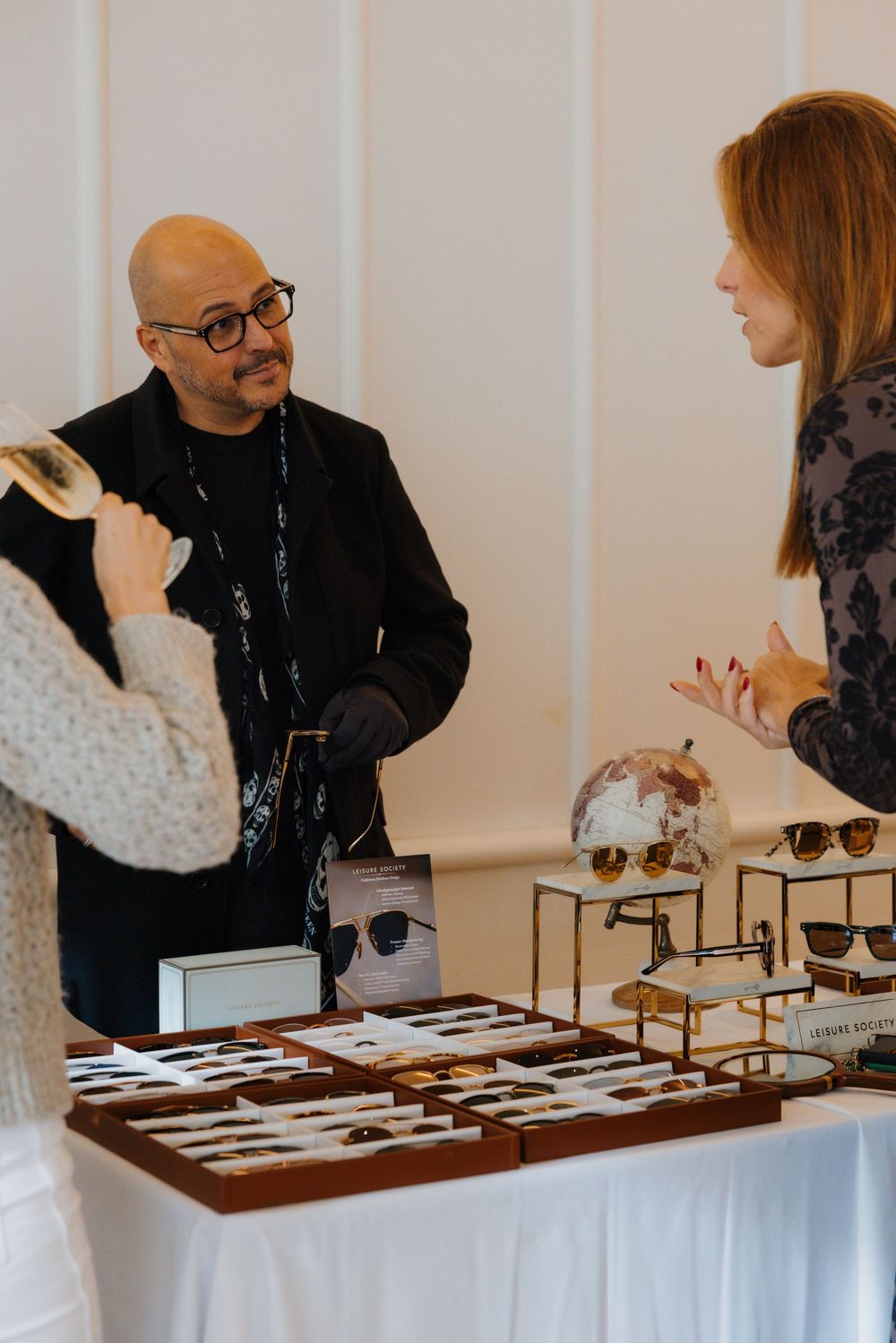 Man in black jacket and glasses stands at a sunglasses display table, conversing with two women at an indoor event.