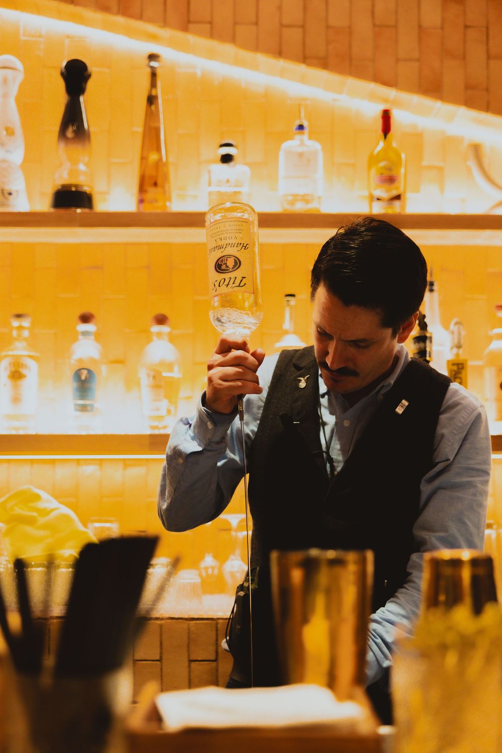 Bartender in vest pouring spirits from a bottle at a warmly lit bar with shelves of liquor bottles.