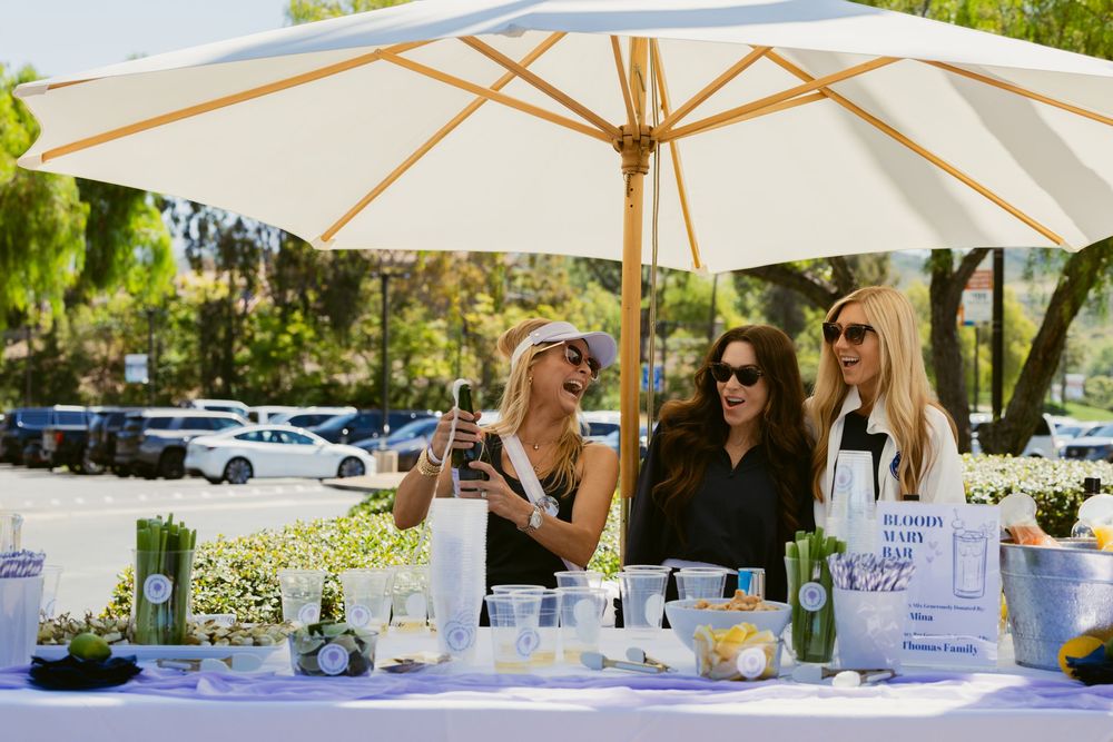 Three women socializing at an outdoor event table with white decor, candles, and greenery under a large umbrella.