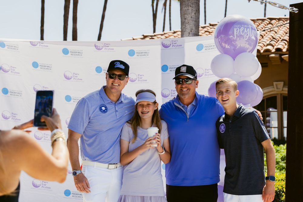 Four people pose for a photo at a charity golf event with purple balloons and a branded backdrop.
