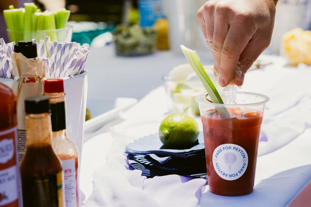 Hand garnishing a Bloody Mary cocktail with celery at an outdoor bar event with hot sauce bottles nearby.
