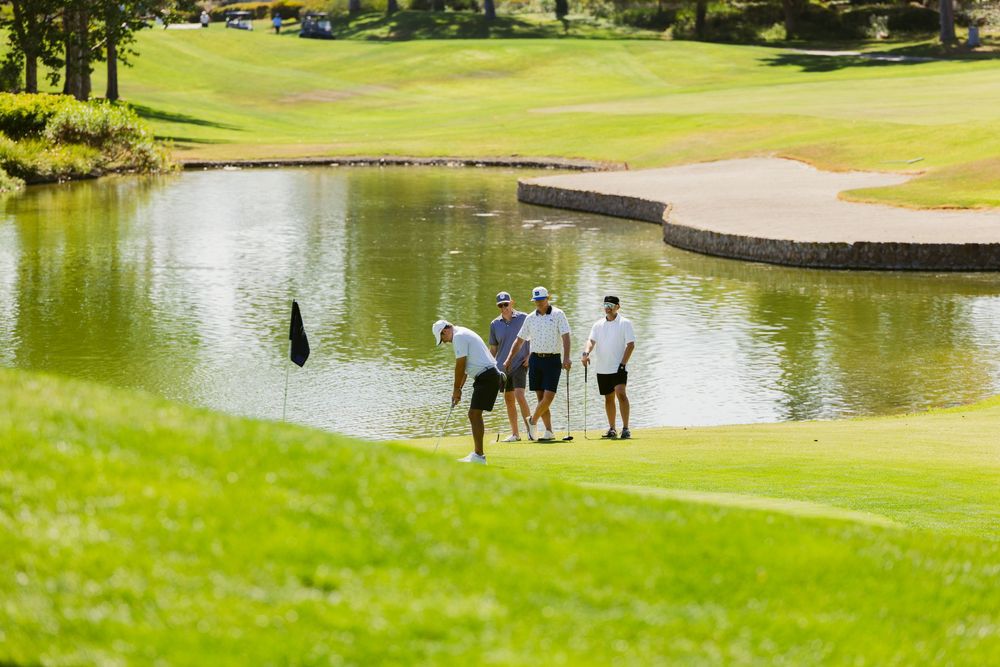Three golfers standing on a lush green golf course beside a serene pond with sand bunkers in the background.