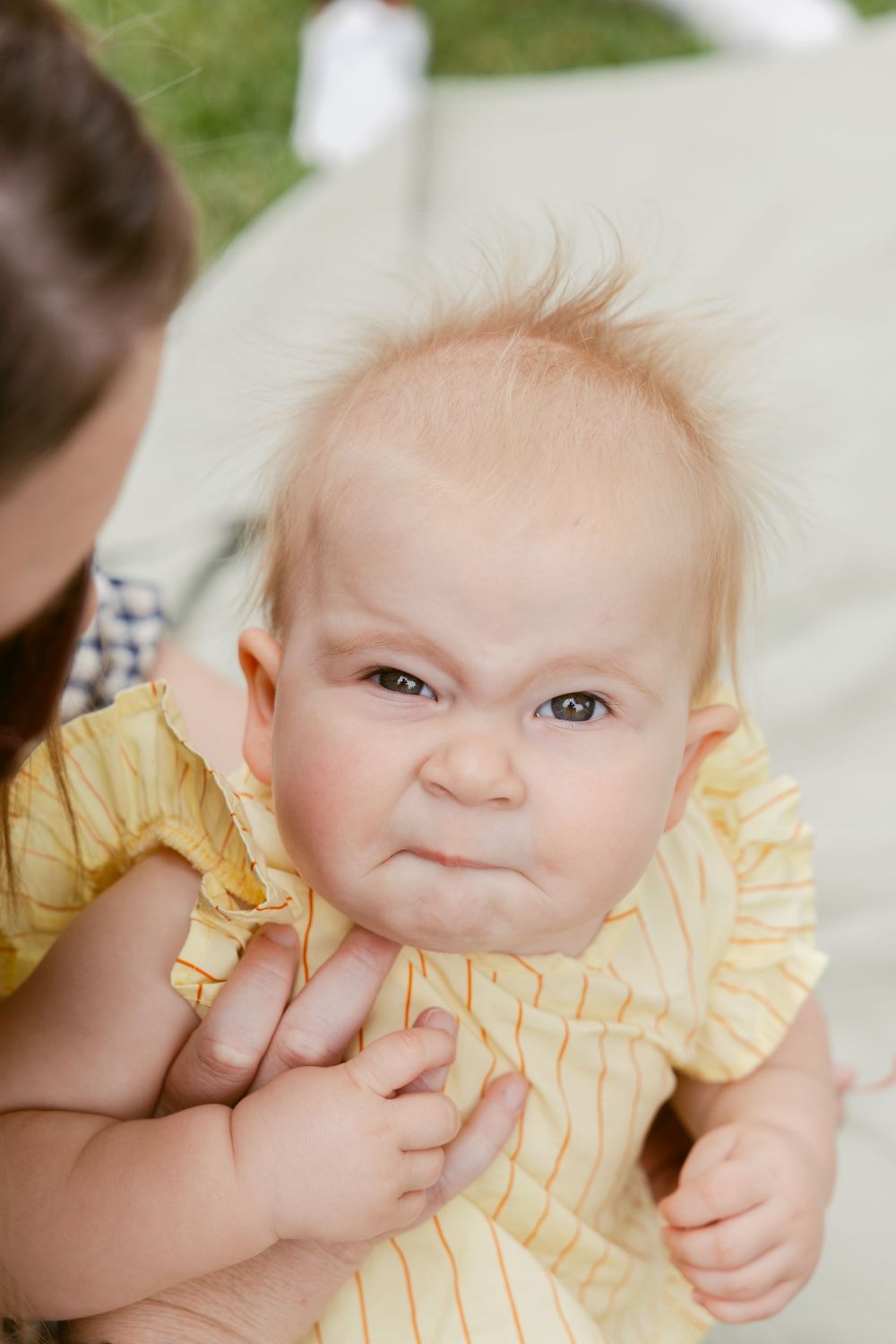 Adorable baby in yellow dress making a grumpy face while being held outdoors.
