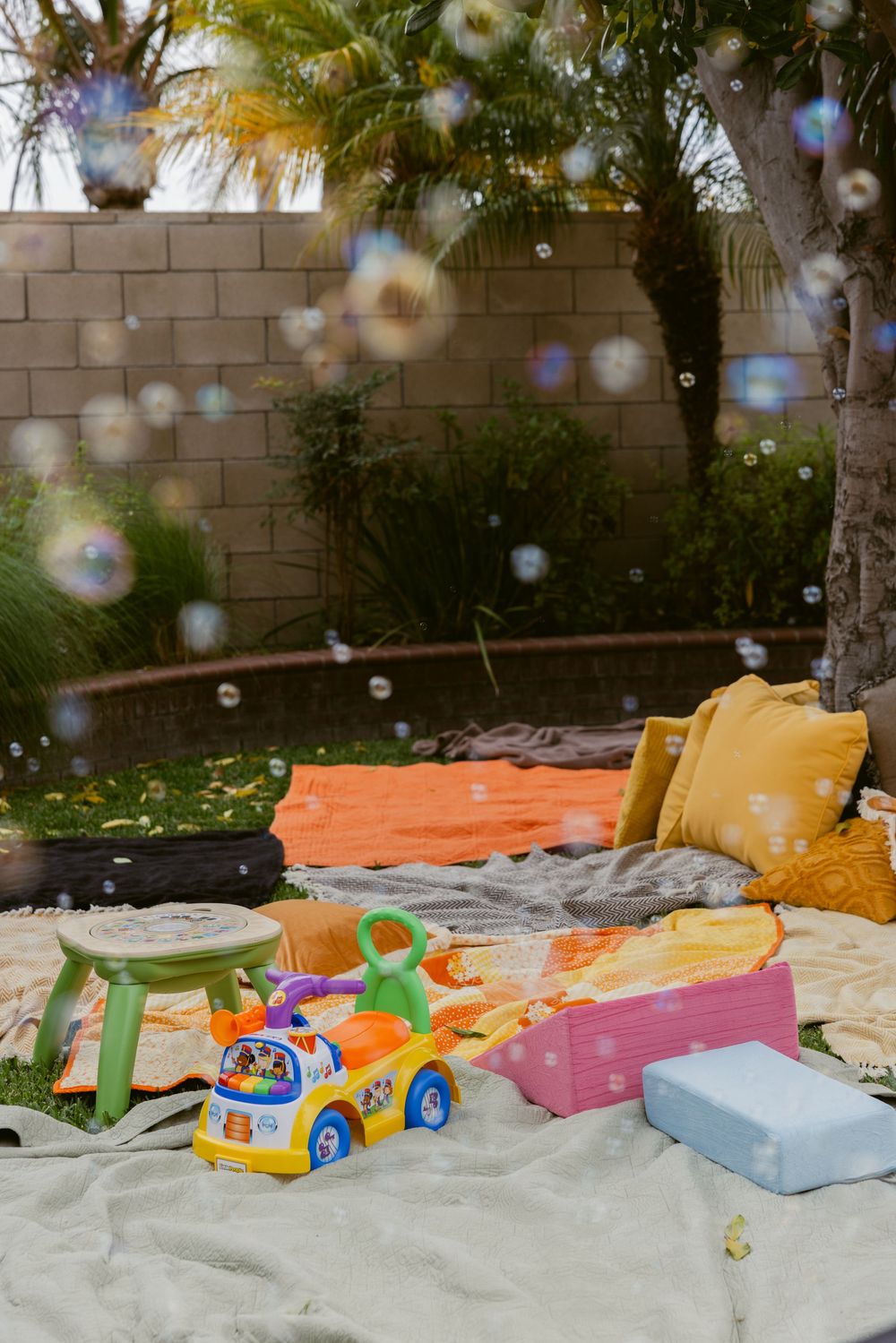 Colorful backyard play area with bubbles, toys, and pillows on blankets near palm trees.