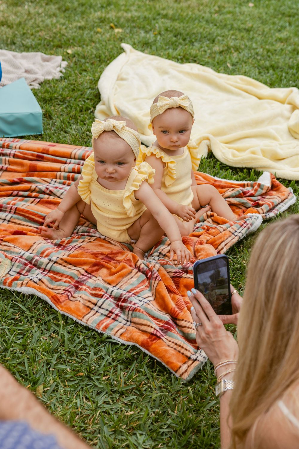 Two baby girls in matching yellow outfits sit on a plaid blanket on grass while a woman photographs them.