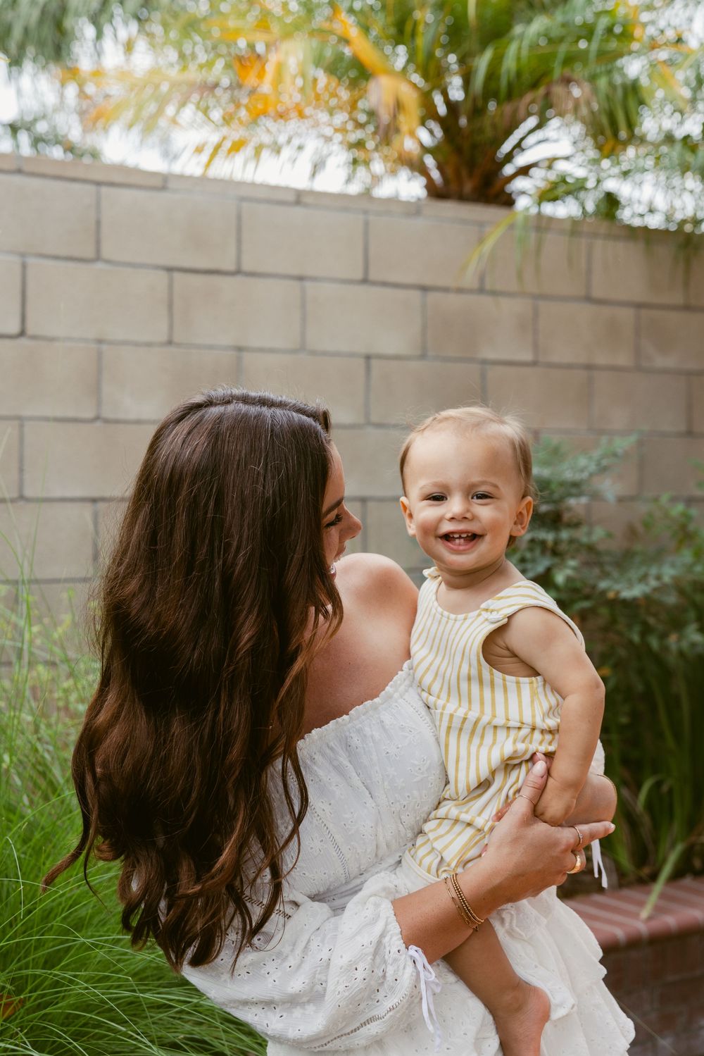 A smiling baby in a yellow striped romper being held by a dark-haired woman in a white dress outdoors.