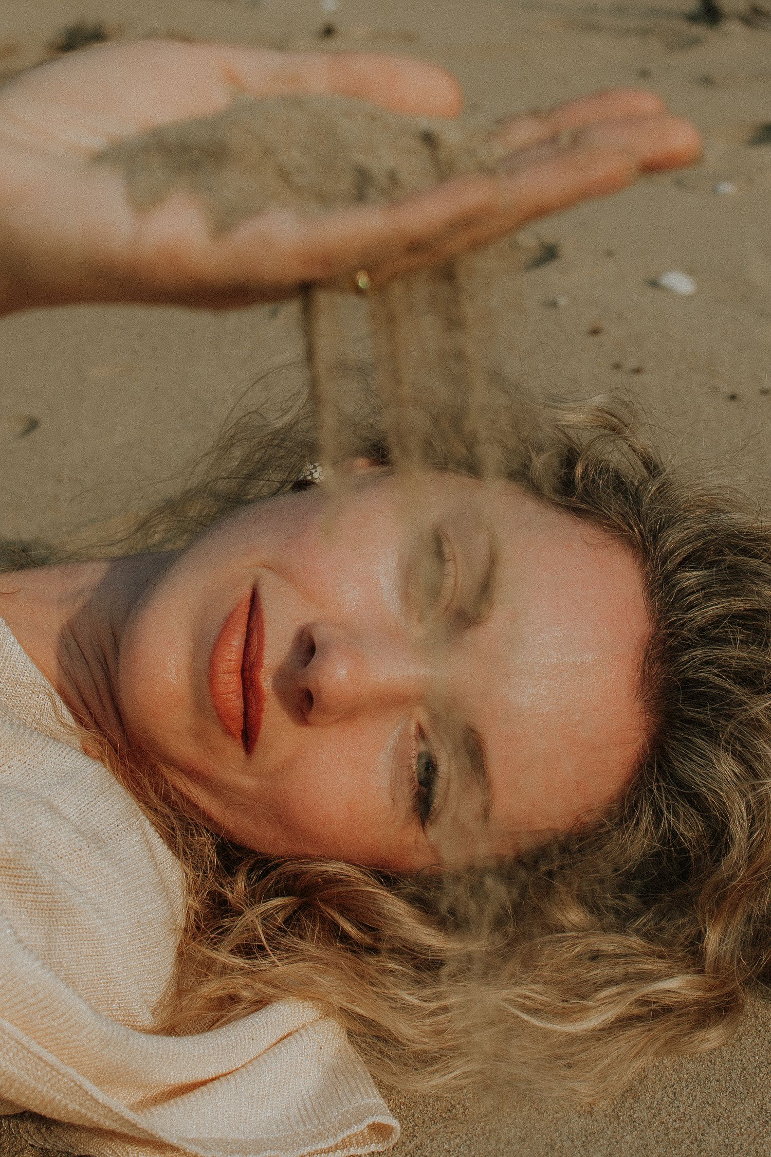 Woman lying on sandy beach with eyes closed, letting sand fall from tattooed hand through fingers.