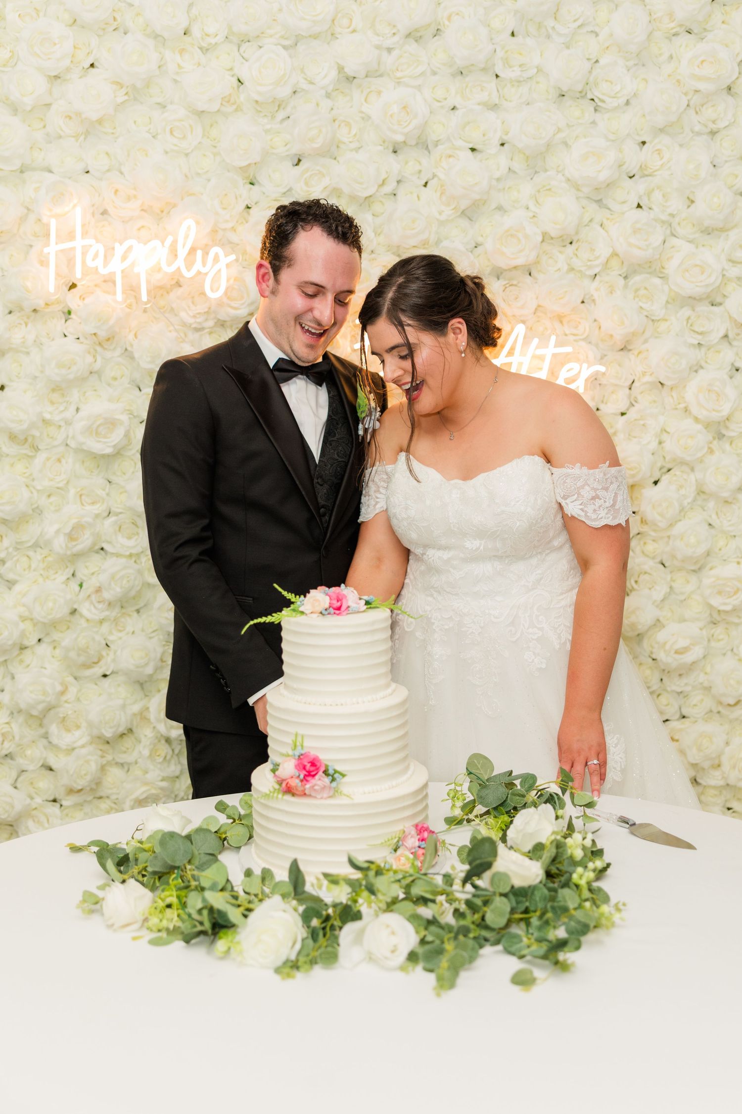 Bride and groom laughing while cutting white tiered wedding cake in front of floral wall with neon sign.