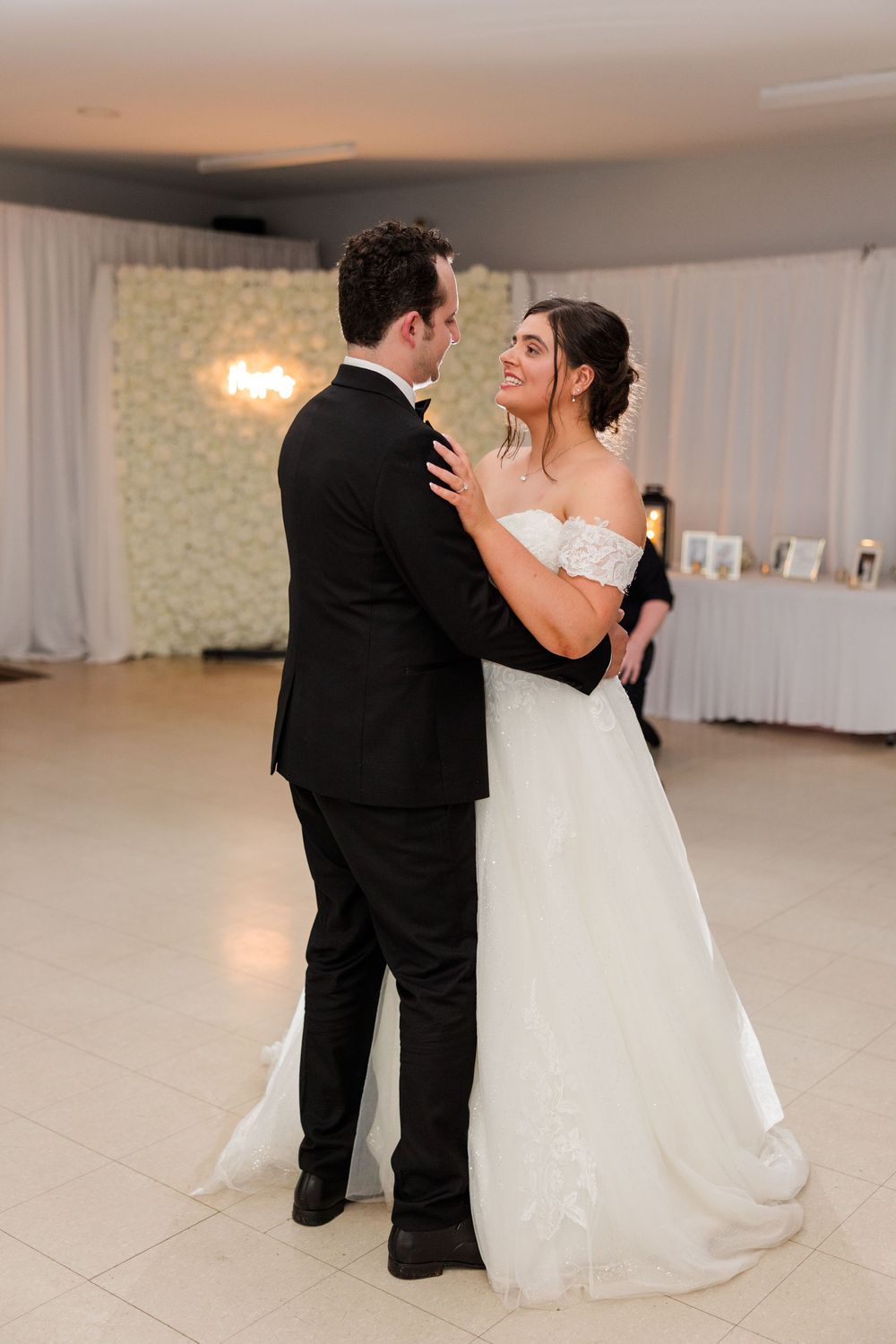 Bride and groom share their first dance at wedding reception, bride in off-shoulder gown smiling at groom in black tux.
