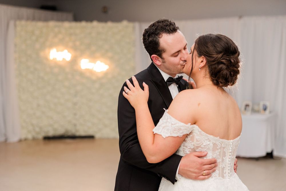 Bride and groom share their first dance kiss, bride in lace off-shoulder gown, groom in black tuxedo, floral wall backdrop.