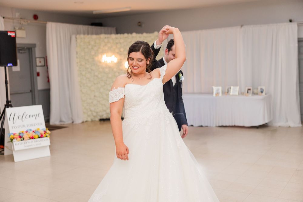 Bride in off-shoulder white ball gown twirling during first dance at elegant wedding reception.