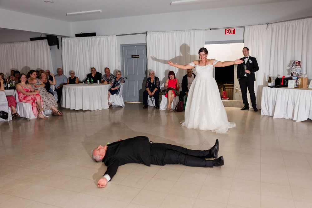 Bride stands with arms outstretched over groom lying on dance floor at wedding reception.
