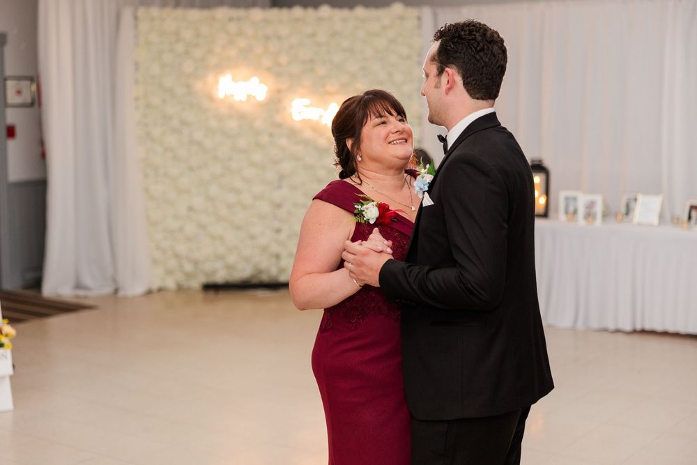 Mother and son share a tender first dance at a wedding reception, she in a red gown, he in a black tuxedo.