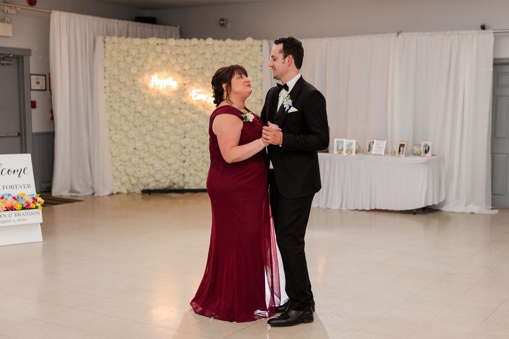 A couple shares a mother-son dance at a wedding reception, with a floral wall backdrop.