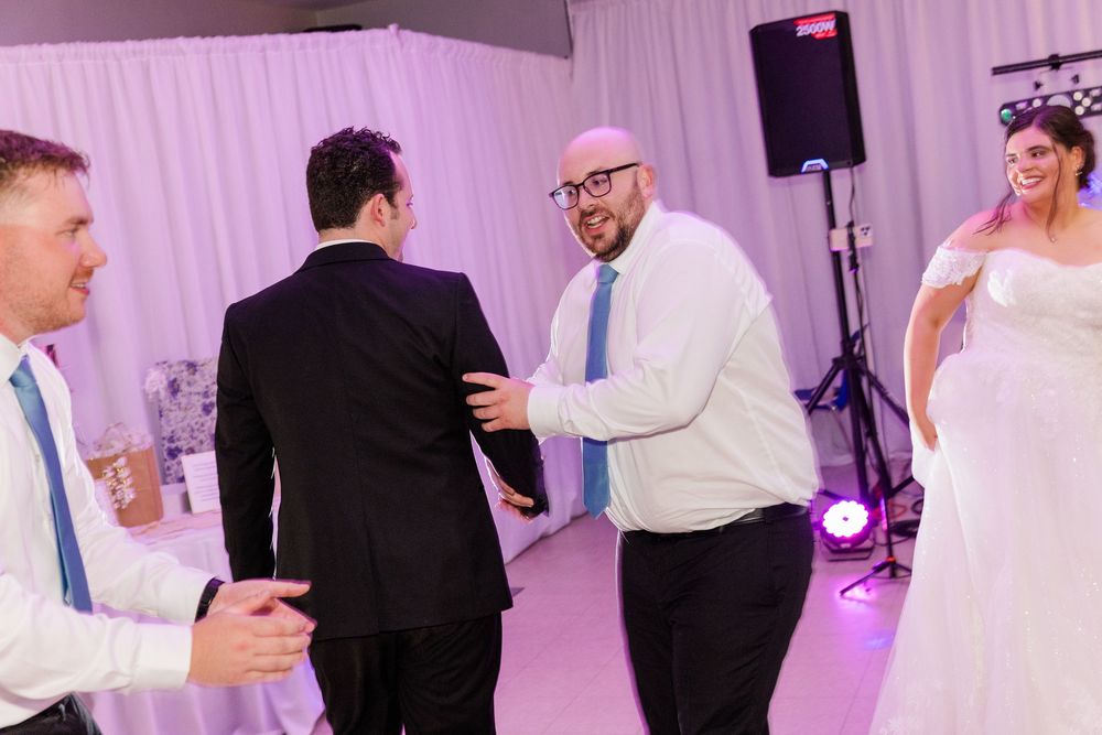 Guests dancing joyfully at a wedding reception, with a smiling bride in a white gown visible in the background.