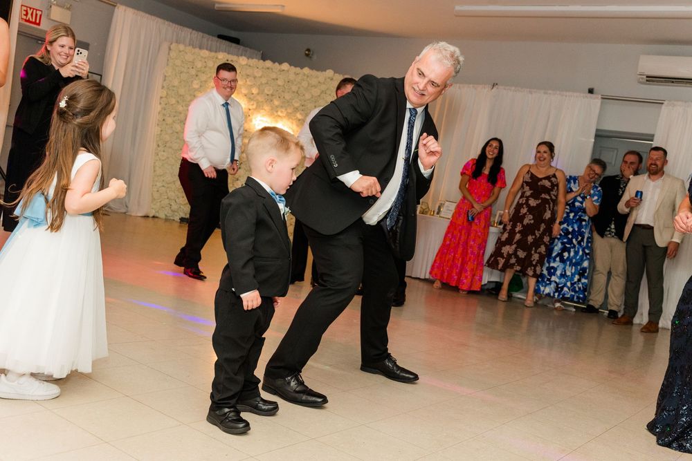 An elderly man dances with a young boy in a suit at a wedding reception while guests watch and celebrate.